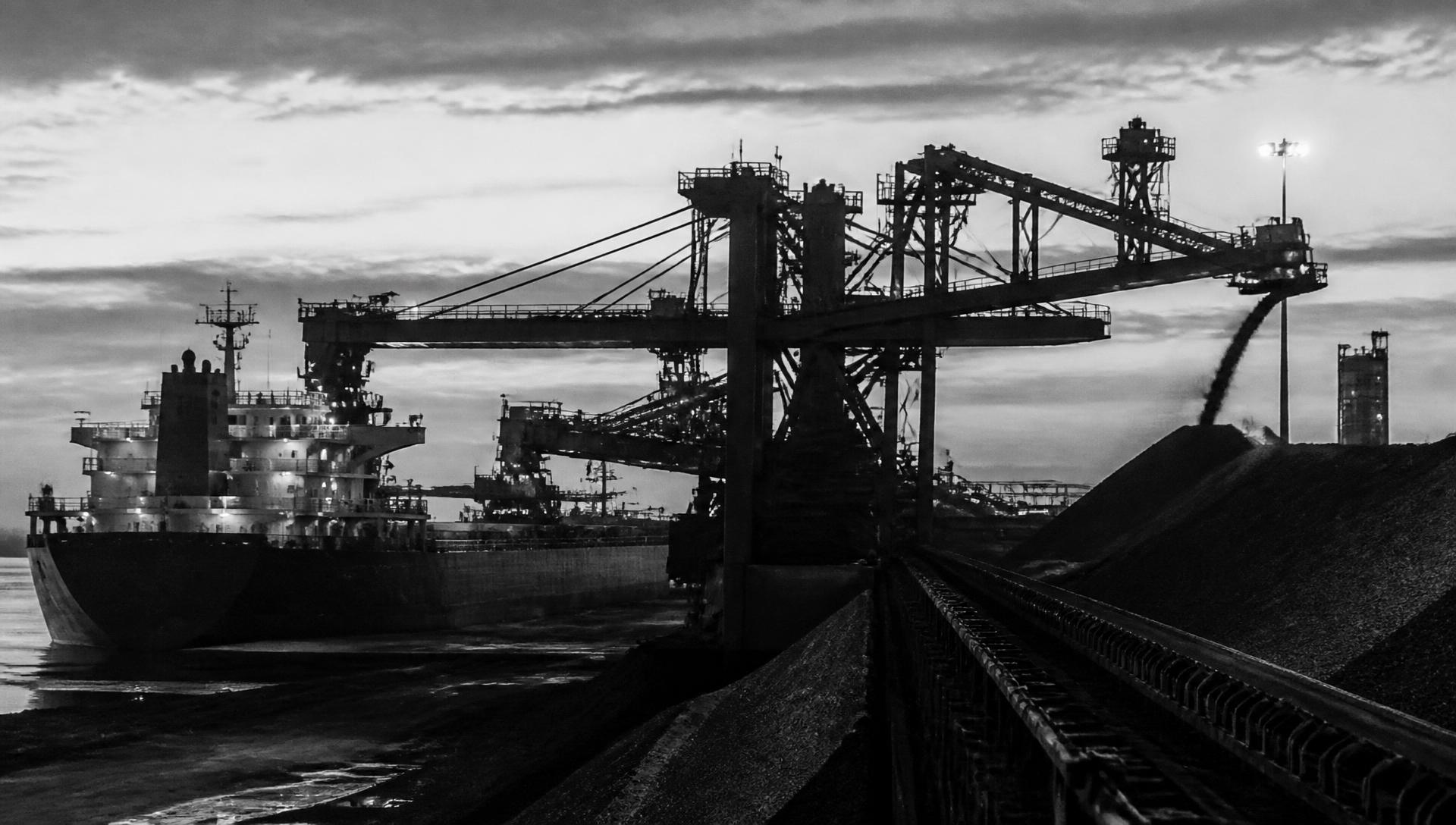 Monochrome photograph of a bulk cargo terminal: ship loaders and conveyor gantries alongside a bulk carrier